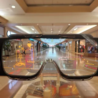 The image captures a first-person perspective of a user experiencing an advanced augmented reality interface through a pair of smart glasses. As the wearer gazes along the interior of a shopping mall, digital overlays provide interactive icons and information, seamlessly integrating with the user's field of vision. This cutting-edge technology blends the physical and digital worlds, creating a dynamic and interactive environment for the user to navigate and engage with.