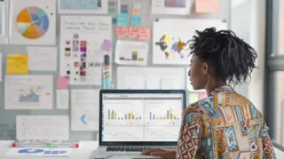 In a contemporary office setting, a professional woman is deeply focused as she analyzes complex data displayed across dual computer screens. The room is well-lit, enhancing the clarity of several informational boards in the background, which are filled with colorful charts, graphs, and notes. Her attention to detail is evident as she possibly searches for trends, insights, or solutions to drive strategic business decisions.