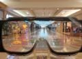 The image captures a first-person perspective of a user experiencing an advanced augmented reality interface through a pair of smart glasses. As the wearer gazes along the interior of a shopping mall, digital overlays provide interactive icons and information, seamlessly integrating with the user's field of vision. This cutting-edge technology blends the physical and digital worlds, creating a dynamic and interactive environment for the user to navigate and engage with.