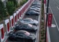 This image captures a modern electric vehicle charging station bustling with activity as multiple cars are docked at the red charging points. The time seems to be twilight, with the sky showcasing beautiful gradients of color, fading from blue to the warmth of the disappearing sun. The neatly aligned cars, with their diverse colors and designs, reflect a commitment to sustainable transportation. In the background, gentle hills paint a serene and natural contrast to the technological advancement represented by the charging station. The scene not only highlights the growth in electric vehicle adoption but also illustrates a harmonious blend of innovation with the environment.