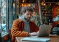 In the calming ambiance of a warmly lit café, a young man sits engrossed in his laptop. The window beside him reveals a hint of the bustling street outside, yet the tranquility within the cafe provides a serene backdrop for work and reflection. His focused gaze, slightly furrowed brow, and the casual attire suggest a fusion of comfort and productivity that's often sought by remote workers everywhere.