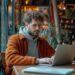 In the calming ambiance of a warmly lit café, a young man sits engrossed in his laptop. The window beside him reveals a hint of the bustling street outside, yet the tranquility within the cafe provides a serene backdrop for work and reflection. His focused gaze, slightly furrowed brow, and the casual attire suggest a fusion of comfort and productivity that's often sought by remote workers everywhere.